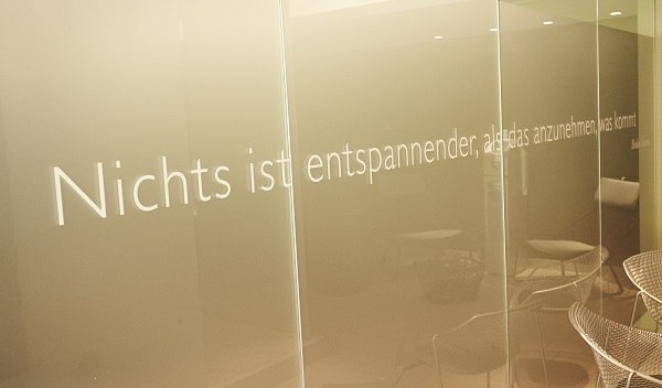 Glaswand mit Zitat: "Nichts ist entspannender, als das anzunehmen, was kommt." Im Hintergrund sind Stühle sichtbar., © Waldhotel Stuttgart GmbH