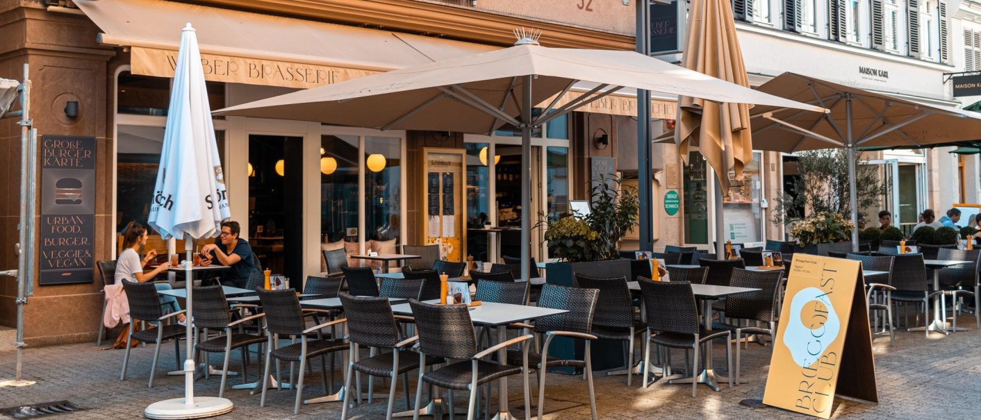 Ein Café mit Terrasse, Tischen und Stühlen, Sonnenschirmen und Gästen. Ein Schild wirbt für Frühstücksangebote., © Stuttgart Marketing GmbH, Sarah Schmid