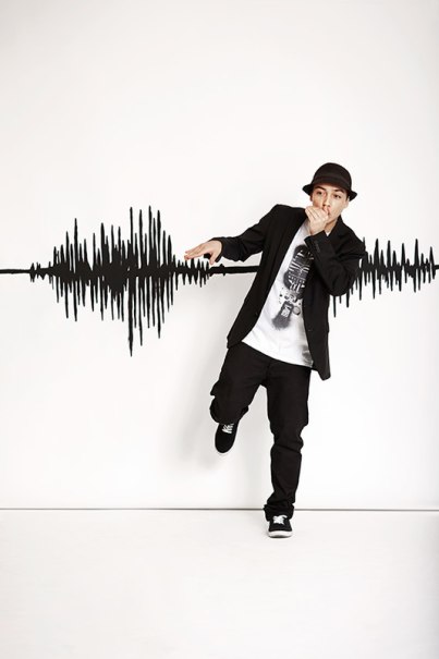 A beatboxer poses in front of a wall with sound wave graphics, wearing a hat and holding his hand in front of his mouth. He is casually dressed in black and white., &copy; Renitenztheater Stuttgart e.V.