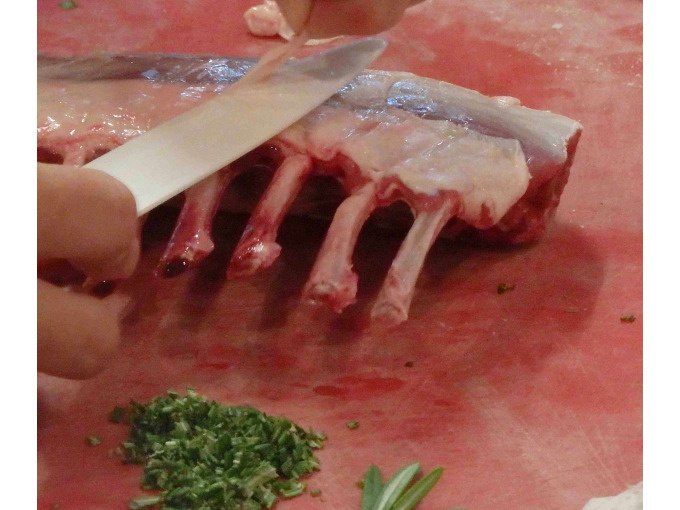 A rack of lamb is cut with a knife on a chopping board. Fresh herbs are placed next to it., &copy; Kultweine-Kochwelten-Kunst&uuml;cke - Kochschule Sonja Lenz