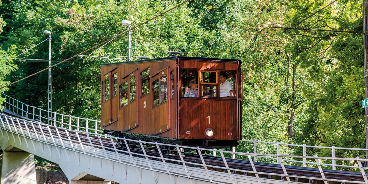 Eine historische Standseilbahn mit Holzverkleidung f&auml;hrt eine geneigte Strecke entlang, umgeben von dichtem Gr&uuml;n., &copy; TMBW, Gregor Lengler