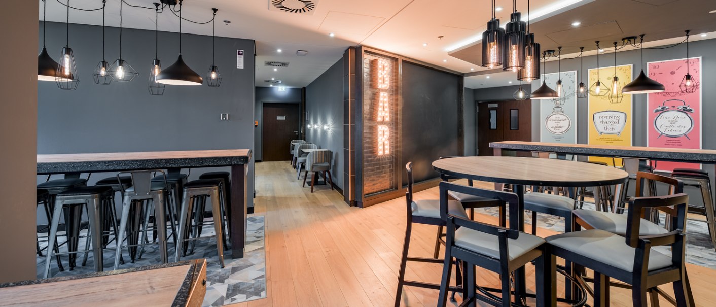 Modern breakfast area with high tables, chairs, stylish lamps and a bright 'BAR' sign on the wall., © Premier Inn Modern breakfast area with high tables, chairs, stylish lamps and a bright 'BAR' sign on the wall., © Premier Inn