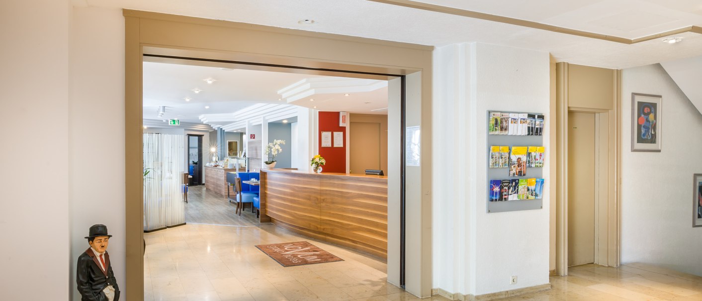 Bright hotel lobby with reception, blue chairs and a brochure rack on the wall. A figure stands in the corner., © TOMAS