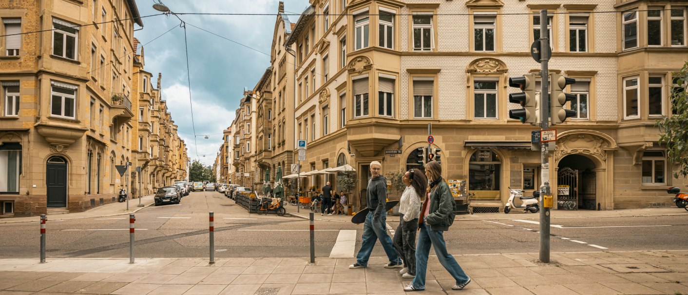 Drei Personen überqueren eine Straße in einer Stadt mit historischen Gebäuden. Der Himmel ist bewölkt, und die Straße ist belebt., © SMG Stuttgart Marketing GmbH - Sarah Schmid Drei Personen überqueren eine Straße in einer Stadt mit historischen Gebäuden. Der Himmel ist bewölkt, und die Straße ist belebt., © SMG Stuttgart Marketing GmbH - Sarah Schmid