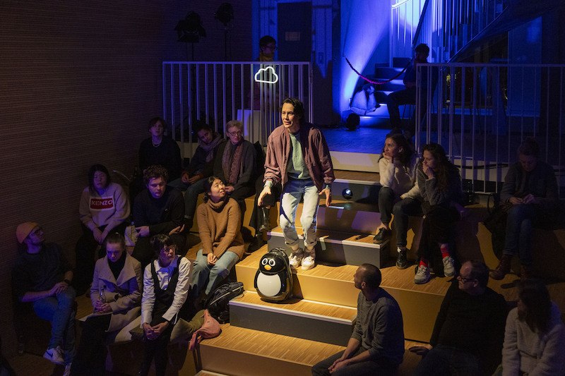 Ein Mann steht in einem kleinen Theater und spricht zu einer Gruppe von Menschen. Die Bühne ist blau beleuchtet., © tri-bühne e.V.