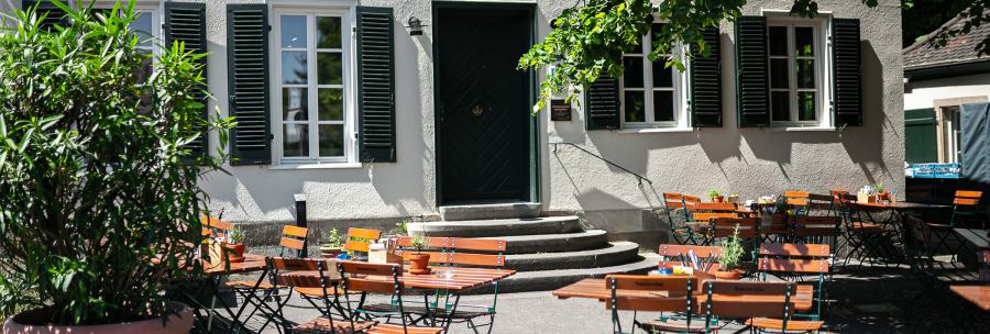 An empty beer garden with wooden tables and chairs in front of a building labeled 'Wirtshaus Hotel'. Shadows from trees fall on the ground., &copy; Cara Schmid