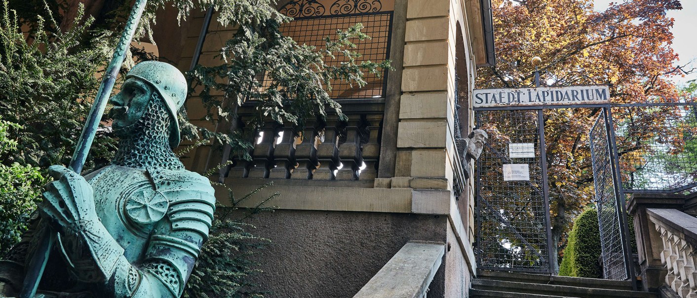Eingang zum Städtischen Lapidarium mit einer grünen Ritterstatue im Vordergrund und einem Tor im Hintergrund., © Städtisches Lapidarium, Foto Julia Ochs