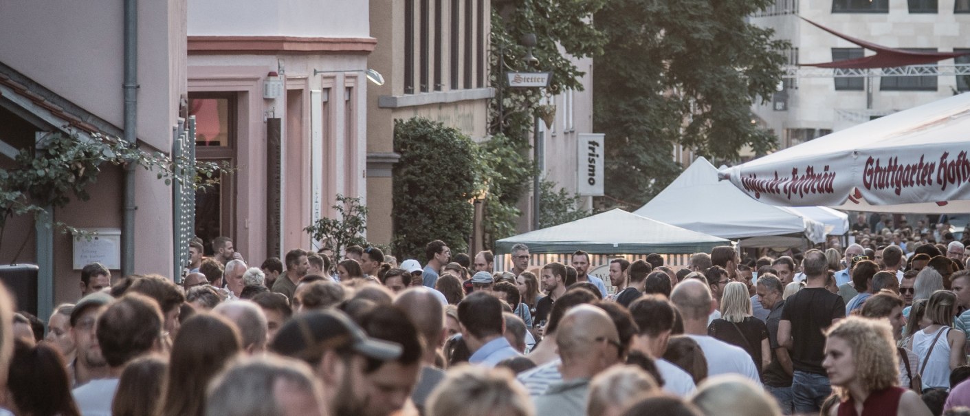 Große Menschenmenge auf einem Straßenfest, umgeben von Gebäuden und Zelten. Im Hintergrund sind Schilder und Bäume zu sehen., © SMG, trickytine Große Menschenmenge auf einem Straßenfest, umgeben von Gebäuden und Zelten. Im Hintergrund sind Schilder und Bäume zu sehen., © SMG, trickytine