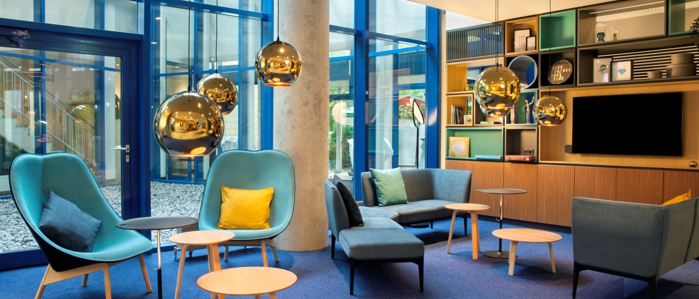 Moderne Lounge mit blauen Sesseln, Holztischen und goldenen H&auml;ngelampen. Gro&szlig;e Fenster und Regale im Hintergrund schaffen eine einladende Atmosph&auml;re., &copy; Holiday Inn Stuttgart
