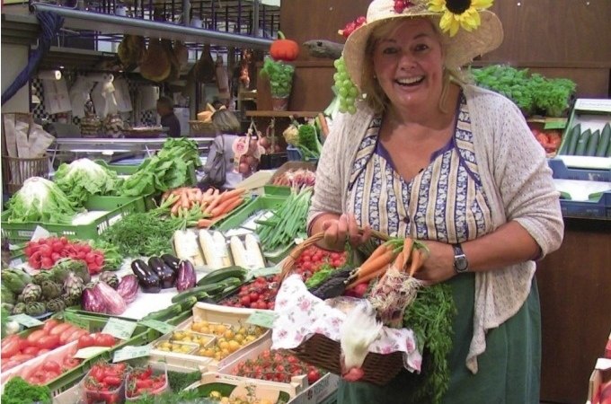 Frau mit Sonnenblumenhut in einer Markthalle, lächelnd, umgeben von frischem Gemüse und Obst. Sie hält einen Korb mit Karotten und Kräutern., © Stuttgart Marketing GmbH