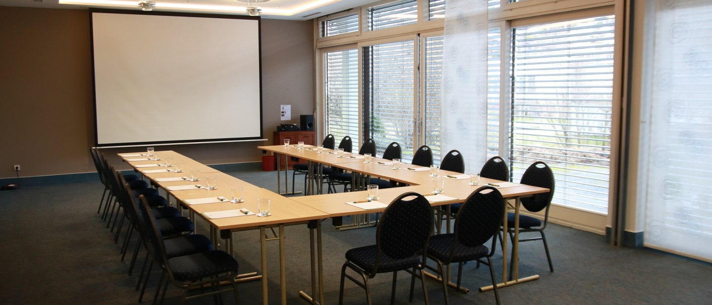 Moderner Tagungsraum mit U-förmiger Tischanordnung, Stühlen, großem Bildschirm und Fenstern mit Jalousien., © ©hotel-graf-eberhard.de
