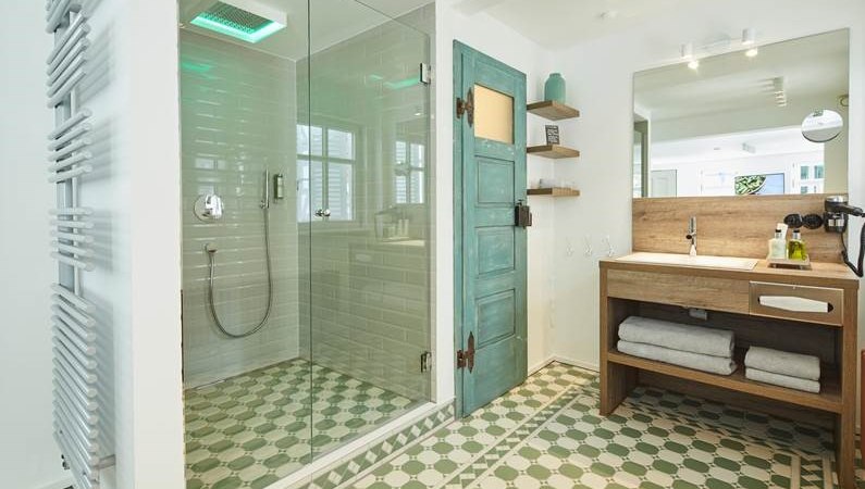 Modernes Badezimmer mit Glasdusche, grünem Fliesenboden, Holzwaschtisch und türkisfarbener Tür. Handtücher und Pflegeprodukte sind bereitgestellt., © Hotel Restaurant Bachofer