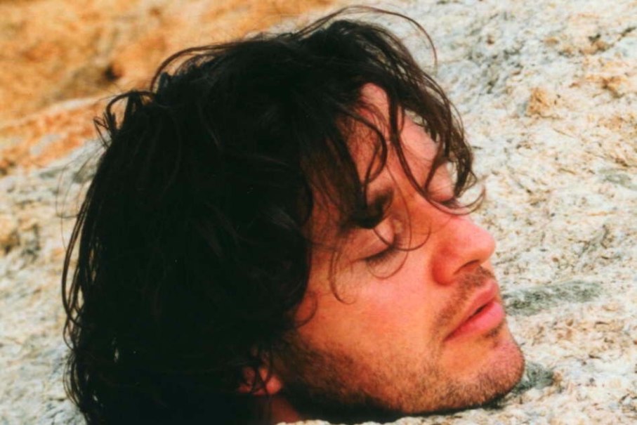 Ein Mann mit dunklem, lockigem Haar und geschlossenen Augen liegt in Sand oder Erde eingebettet. Sein Gesicht wirkt entspannt., © kaba-reh production Ein Mann mit dunklem, lockigem Haar und geschlossenen Augen liegt in Sand oder Erde eingebettet. Sein Gesicht wirkt entspannt., © kaba-reh production