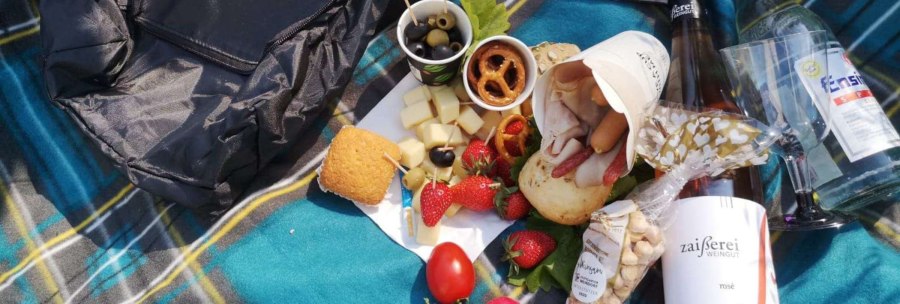 A picnic on a checkered blanket with cheese, olives, strawberries, bread, wine and a bottle of water. A rucksack lies next to it., © Weingut Zaißerei