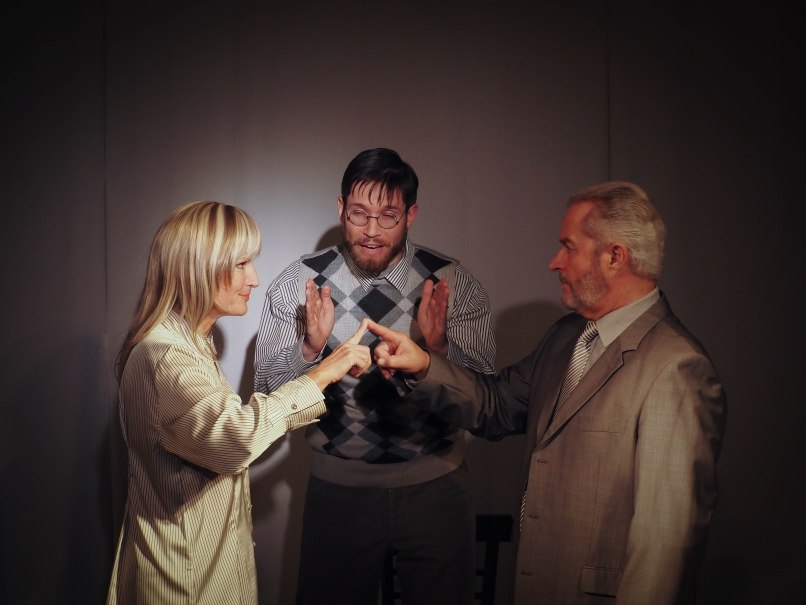 Drei Personen in einer Theaterszene. Zwei zeigen mit Fingern aufeinander, während die dritte Person dazwischen steht und gestikuliert., © Theater Atelier