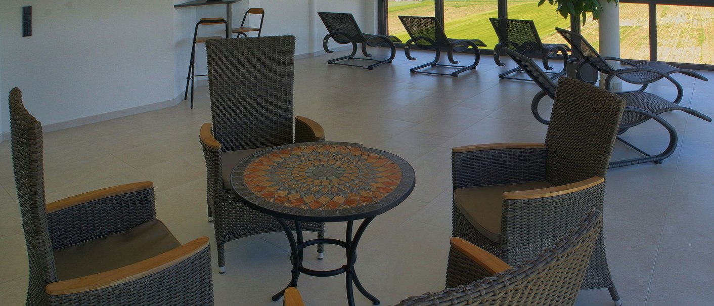 Relaxation room with rattan furniture and loungers. Large windows offer a view of the green landscape. A table with a mosaic pattern stands in the foreground., © TOMAS