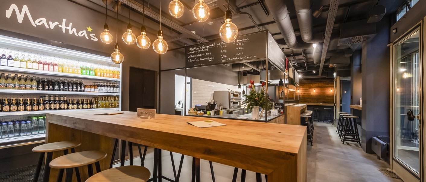 Modernes Café mit Holztischen, Getränkekühlschrank und hängenden Glühbirnen. An der Wand steht "Martha's". Gemütliche Atmosphäre mit Barbereich., © Martha's, Stuttgart