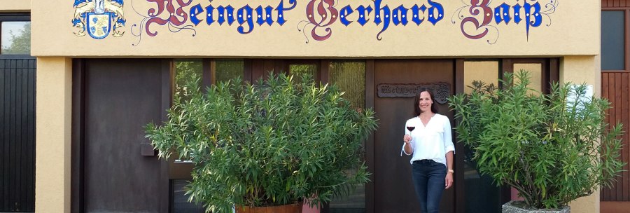 Eine Frau steht vor dem Weingut Gerhard Zaiß, hält ein Glas Rotwein und lächelt. Das Gebäude hat eine dekorative Beschriftung und Pflanzen im Vordergrund., © Simone Mack Eine Frau steht vor dem Weingut Gerhard Zaiß, hält ein Glas Rotwein und lächelt. Das Gebäude hat eine dekorative Beschriftung und Pflanzen im Vordergrund., © Simone Mack