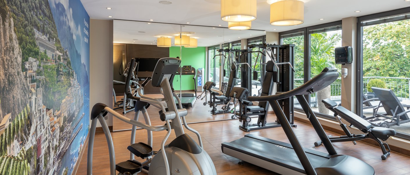 Modernes Fitnessstudio mit Laufband, Crosstrainer und Kraftgeräten. Große Fenster bieten Blick ins Grüne, Wandbild zeigt Küstenlandschaft., © NH Hotel Stuttgart Airport