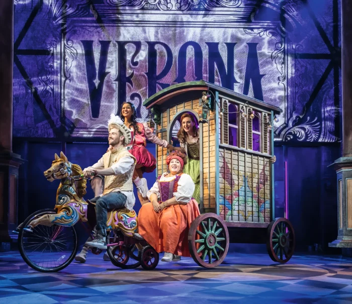 A theater scene featuring a man on a decorative horse-drawn bicycle and three women in a colorful carriage. In the background, a large sign reads "Verona.", © Johan Persson / Stage Entertainment A theater scene featuring a man on a decorative horse-drawn bicycle and three women in a colorful carriage. In the background, a large sign reads "Verona.", © Johan Persson / Stage Entertainment