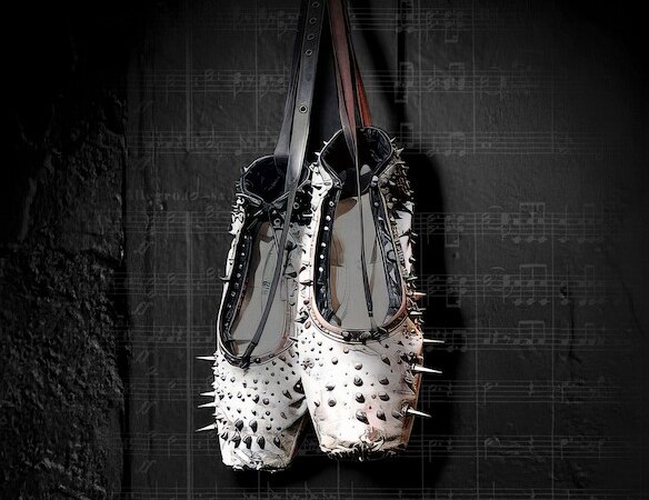 Ein Paar Ballettschuhe mit Nieten h&auml;ngt vor einem dunklen Hintergrund mit Notenbl&auml;ttern. Die Schuhe kombinieren klassische Eleganz mit einem rebellischen Touch., &copy; Theaterhaus Stuttgart e.V.