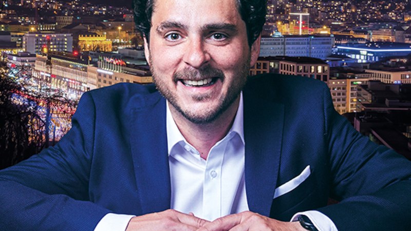 Ein Mann in einem Anzug l&auml;chelt vor einer beleuchteten Stadt bei Nacht. Im Hintergrund sind Lichter und Geb&auml;ude zu sehen., &copy; Altes Schauspielhaus und Kom&ouml;die im Marquardt e.V.