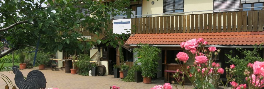 Ein Weinhof mit Holzbalkon, roten Dachziegeln und blühenden Rosen im Vordergrund. Ein Schild mit der Aufschrift 'Weinhof Württemberg Helmut Zaiß' ist sichtbar., © Helmut Zaiß Weingut & Weingastronomie