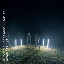 Zwei Personen stehen nachts auf einer Wiese, umgeben von leuchtenden St&auml;ben. Die Szene wirkt mystisch und futuristisch., &copy; links im Bild