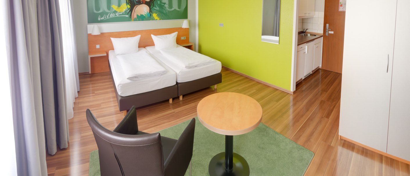 Modernes Superior Doppelzimmer mit zwei Betten, grünem Wandbild, Tisch, Stuhl und kleiner Küchenzeile auf Holzboden., © attimo Hotel Stuttgart Modernes Superior Doppelzimmer mit zwei Betten, grünem Wandbild, Tisch, Stuhl und kleiner Küchenzeile auf Holzboden., © attimo Hotel Stuttgart