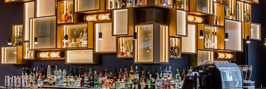 Eine stilvolle Bar mit beleuchteten Regalen voller Flaschen, modernen Barhockern und einer eleganten Theke., © TOMAS Eine stilvolle Bar mit beleuchteten Regalen voller Flaschen, modernen Barhockern und einer eleganten Theke., © TOMAS