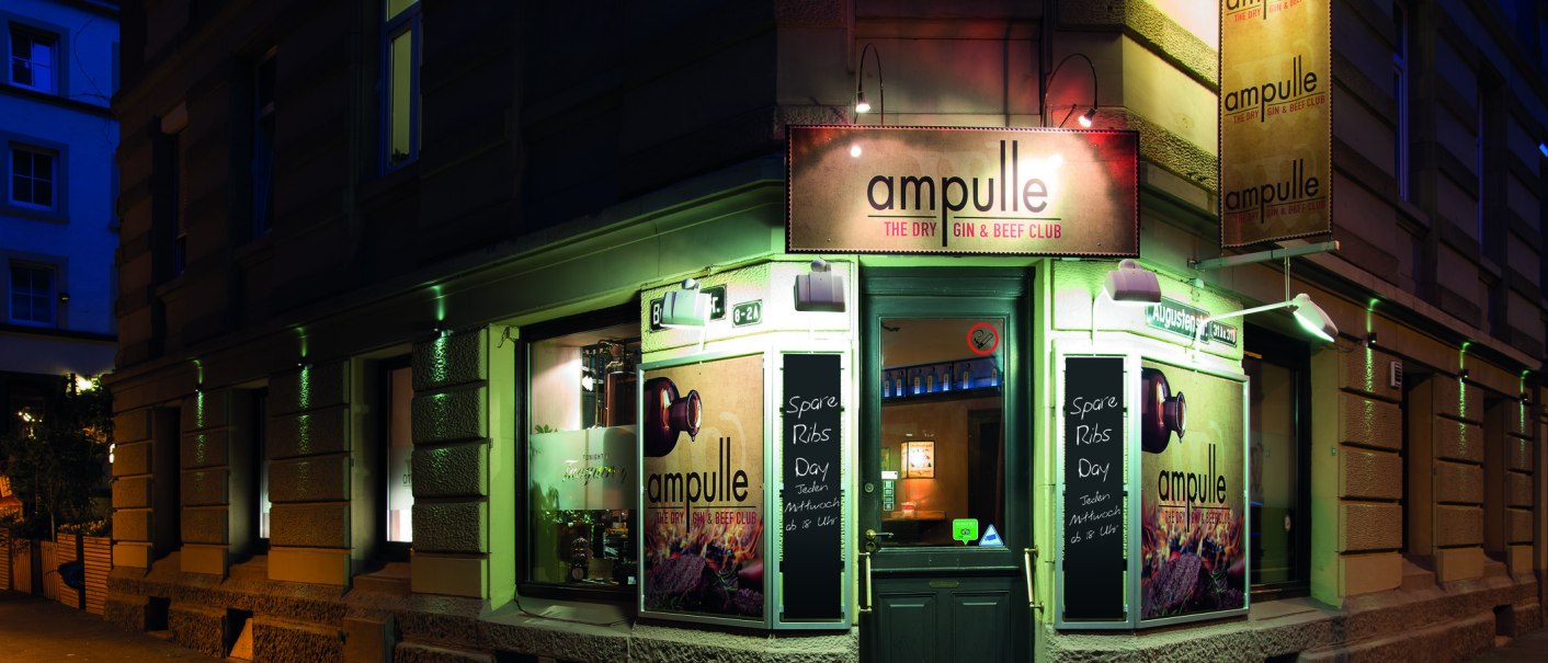 Eingang des Ampulle Gin & Beef Clubs bei Nacht, beleuchtetes Schild und Werbetafeln für Spare Ribs Day. Gebäude an einer Straßenecke., © © Buschwerk Mediendesign-Wilhelmer Gastronomie