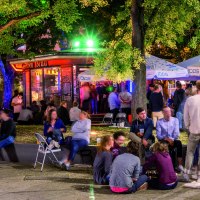 Lively outdoor area with people under illuminated umbrellas and trees. Some are sitting on chairs, others on the ground. Colorful lights in the background., &copy; TMBW, Gregor Lengler