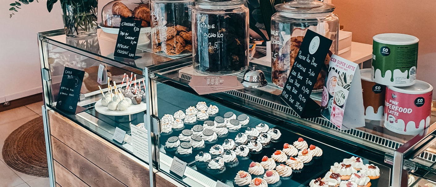 Eine Vitrine in einem Caf&eacute; zeigt eine Auswahl an Cupcakes, Cake Pops und Geb&auml;ck. Daneben stehen Gl&auml;ser mit Keksen und Preisschilder., &copy; Cupcakes & Bagels, Stuttgart