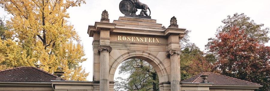 Historic gate with 'Rosenstein' lettering, flanked by buildings. Autumnal trees in the background, two people walking., © TMBW, Christoph Düpper Historic gate with 'Rosenstein' lettering, flanked by buildings. Autumnal trees in the background, two people walking., © TMBW, Christoph Düpper