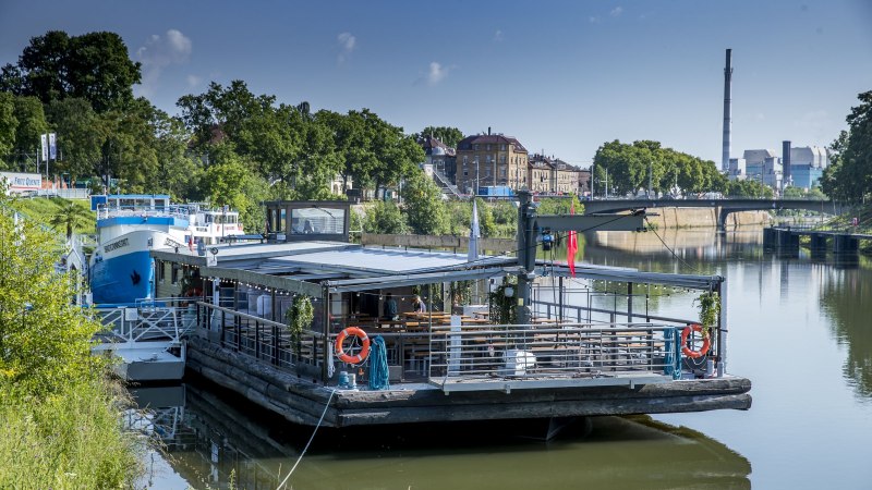 Ein schwimmendes Restaurant auf dem Neckar, umgeben von gr&uuml;ner Vegetation und st&auml;dtischen Geb&auml;uden im Hintergrund., &copy; all copyrights are reserved by maks richter
