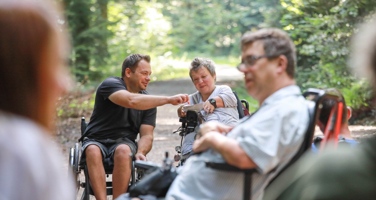 Eine Gruppe von Menschen in Rollst&uuml;hlen unterh&auml;lt sich auf einem Waldweg. Die Umgebung ist gr&uuml;n und sonnig., &copy; NPSFW