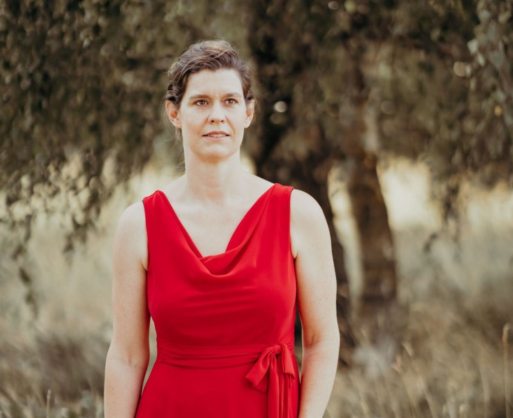 Eine Frau in einem roten Kleid steht im Freien vor einem unscharfen Hintergrund mit Bäumen., © Judith Wiesebrock