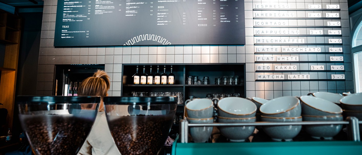 Café-Innenansicht mit Kaffeebohnenbehältern im Vordergrund und Getränkekarte an der Wand. Eine Person steht hinter der Theke., © Stuttgart-Marketing GmbH, Sarah Schmid