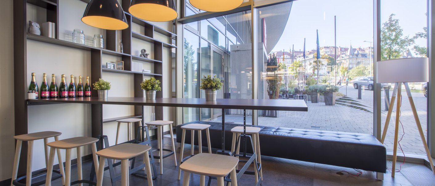 Moderne Lobby mit hohen Tischen, Hockern und großen Lampen. Im Hintergrund sind Regale mit Dekoration und eine Fensterfront mit Blick nach draußen., © Park Inn by Radisson Stuttgart