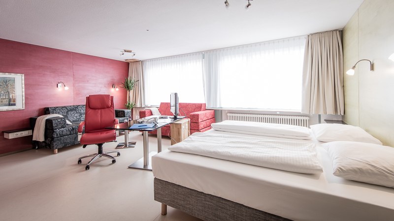 Helles Hotelzimmer mit Doppelbett, Schreibtisch, rotem Sessel und Sofa. Die Wände sind in Rot und Grün gehalten, große Fenster lassen viel Licht herein., © Abalon Hotel Ideal Helles Hotelzimmer mit Doppelbett, Schreibtisch, rotem Sessel und Sofa. Die Wände sind in Rot und Grün gehalten, große Fenster lassen viel Licht herein., © Abalon Hotel Ideal