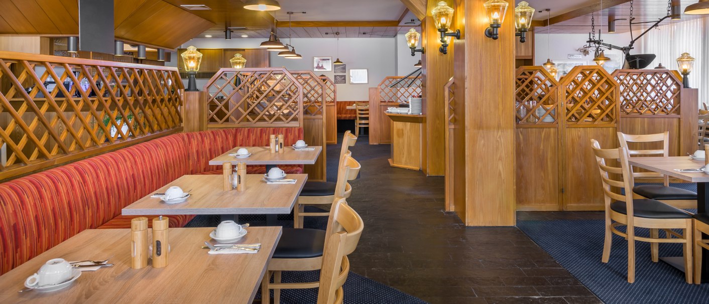 Gemütlicher Frühstücksbereich mit Holzmöbeln, roten Polstern und gedeckten Tischen. Warme Beleuchtung sorgt für eine einladende Atmosphäre., © Novum Management GmbH Gemütlicher Frühstücksbereich mit Holzmöbeln, roten Polstern und gedeckten Tischen. Warme Beleuchtung sorgt für eine einladende Atmosphäre., © Novum Management GmbH