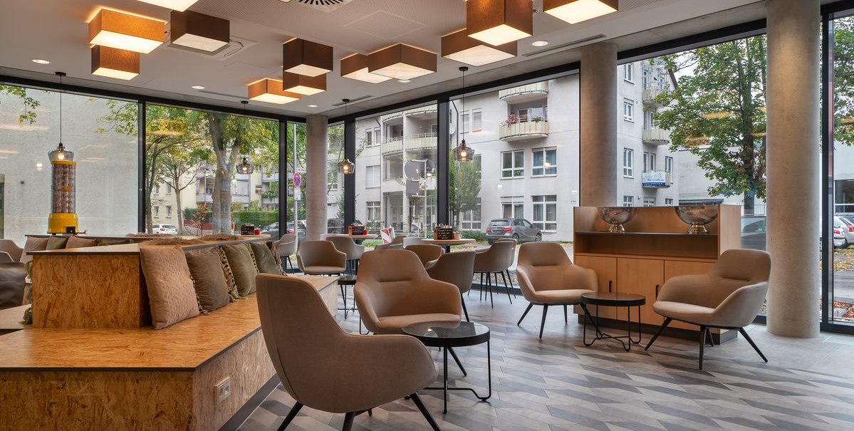 Moderne Lobby mit bequemen Sesseln, Tischen und gro&szlig;en Fenstern. Geometrische Deckenleuchten und ein Blick auf die Stra&szlig;e im Hintergrund., &copy; Novum Hospitality