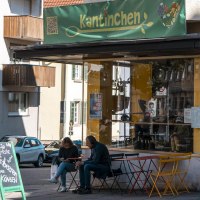 Ein Café namens Kantinchen mit gelbem und grünem Schild. Draußen sitzen Menschen an bunten Tischen. Verkehrsschilder und Autos sind zu sehen., © SMG, Sarah Schmid Ein Café namens Kantinchen mit gelbem und grünem Schild. Draußen sitzen Menschen an bunten Tischen. Verkehrsschilder und Autos sind zu sehen., © SMG, Sarah Schmid