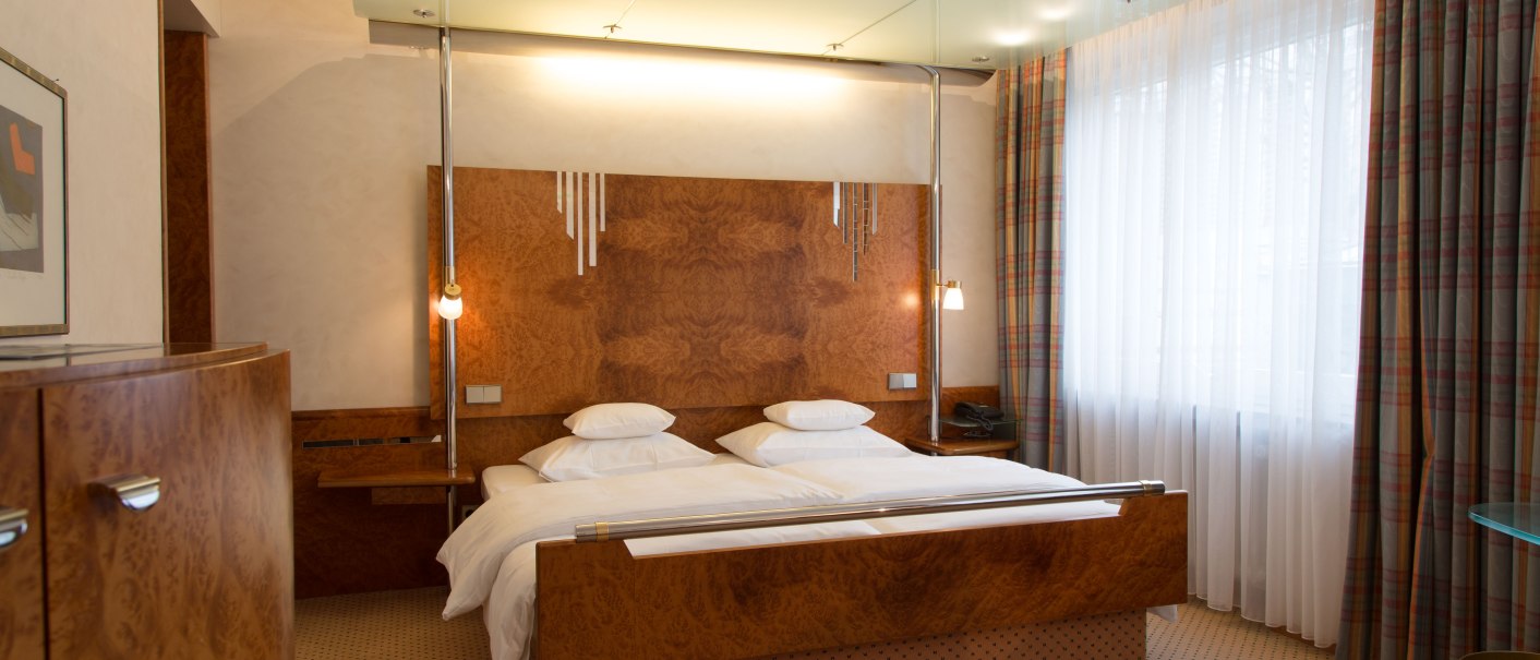 An elegant hotel room with a double bed, wooden furniture, and large windows with curtains. A plate of fruit sits on a glass table., &copy; Hotel Royal