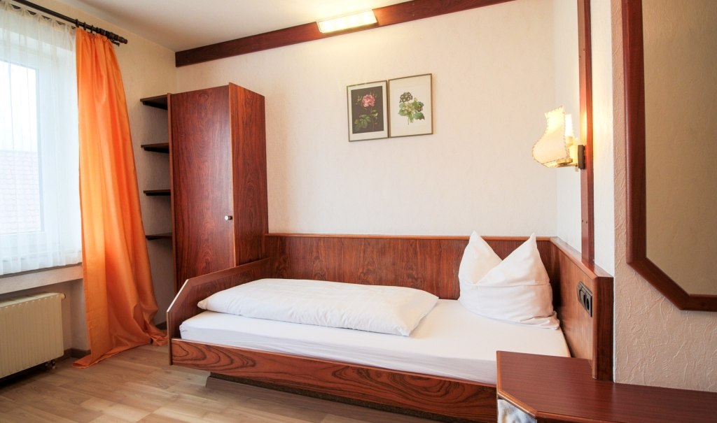 Ein Einzelzimmer mit Holzmöbeln, Einzelbett, orange Vorhängen und Wandbildern. Gemütliche Beleuchtung und helle Wände schaffen eine einladende Atmosphäre., © Ulli Kellenbenz Ein Einzelzimmer mit Holzmöbeln, Einzelbett, orange Vorhängen und Wandbildern. Gemütliche Beleuchtung und helle Wände schaffen eine einladende Atmosphäre., © Ulli Kellenbenz