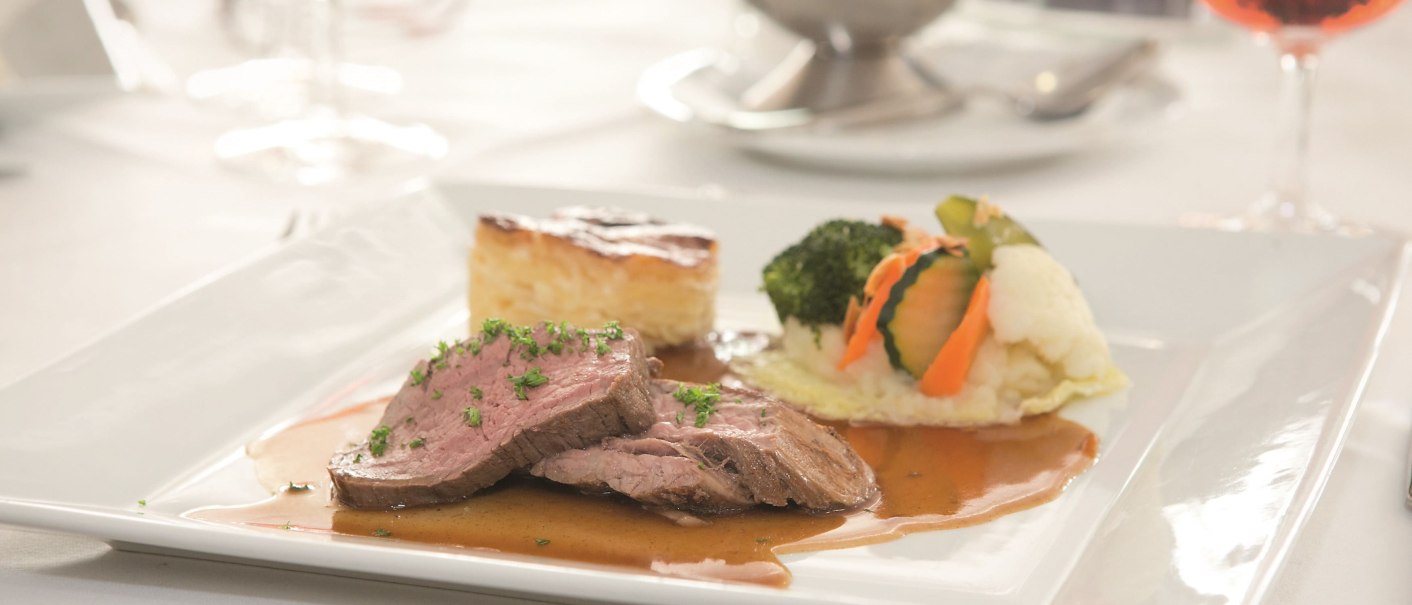 An elegant plate of meat, vegetables and potato gratin, served on a white table with a wine glass in the background., © Erikson Hotel