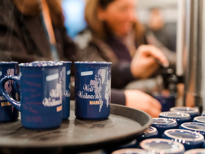 Blaue Tassen mit 'Welt Weihnachts Markt' stehen auf einem Tablett. Im Hintergrund schenken Menschen Glühwein ein, was eine festliche Atmosphäre schafft., © Cool-Tours StattReisen Blaue Tassen mit 'Welt Weihnachts Markt' stehen auf einem Tablett. Im Hintergrund schenken Menschen Glühwein ein, was eine festliche Atmosphäre schafft., © Cool-Tours StattReisen