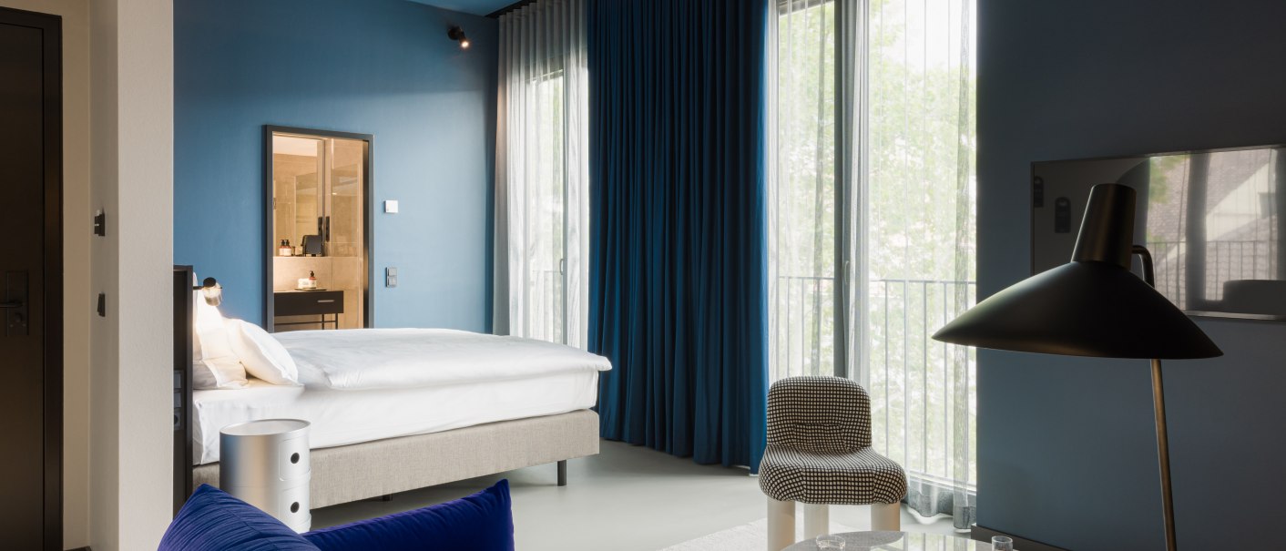 Modernes Hotelzimmer mit blauem Sofa, Bett, Tisch und Lampe. Große Fenster mit blauen Vorhängen und hellem Teppich., © EmiLu Hotel GmbH