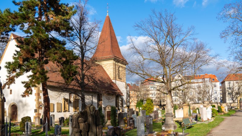The Uff Cemetery, with its church, gravestones, and trees under a blue sky; residential buildings can be seen in the background., © Peter Jagusch/Landeshauptstadt Stuttgart The Uff Cemetery, with its church, gravestones, and trees under a blue sky; residential buildings can be seen in the background., © Peter Jagusch/Landeshauptstadt Stuttgart