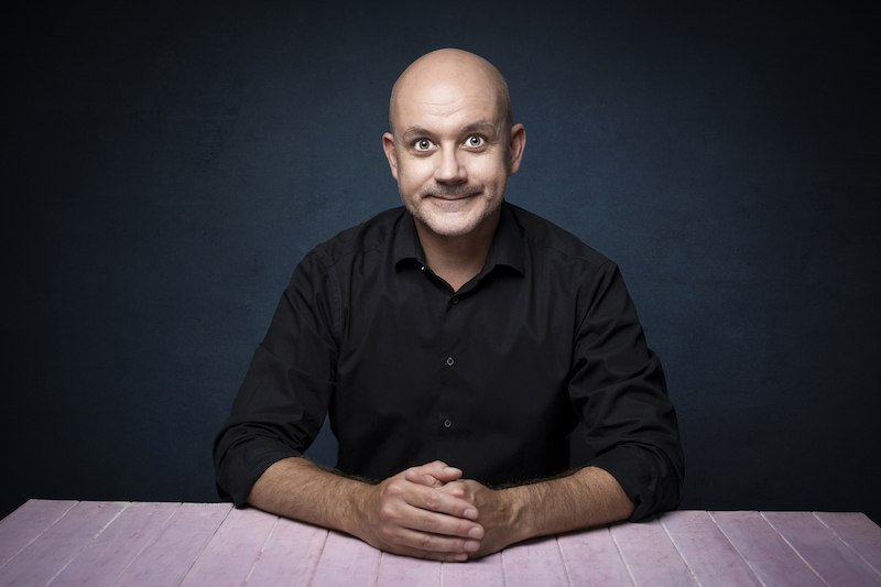 Ein Mann in schwarzem Hemd sitzt l&auml;chelnd an einem rosa Tisch vor einem dunklen Hintergrund., &copy; Theaterhaus Stuttgart e.V.